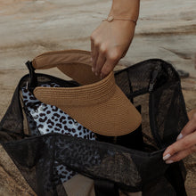 Load image into Gallery viewer, A person places a brown visor into a Thandana Large Black Mesh Beach Bag, with a leopard print waterproof pouch inside. Made from water-resistant fabric, it's ideal for the beach. Set against a stone or concrete background, the person wears a delicate bracelet.