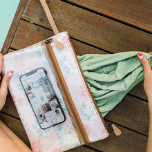 Load image into Gallery viewer, Someone places a Sticky Fingers Clear Pouch Laminated Fabric in Dazzling Blue by Thandana onto a wooden surface. Inside, a smartphone displays images within the clear floral-patterned pouch featuring a zipper and brown strap. The setting near the pool emphasizes the water-resistant qualities.