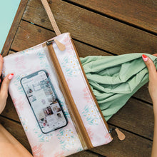 Load image into Gallery viewer, A person with pink nail polish places a green garment into a Sticky Fingers Clear Pouch Laminated Fabric - Jet by Thandana on a wooden table. A smartphone inside the water-resistant pouch displays a social media app.