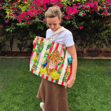 Load image into Gallery viewer, Marzelle holding the  Lush Leopard Jungle Beach Bag by Natural Selection in her garden with pink flowers in the background. The large beach bag features a leopard surrounded by green banana leaves, bananas and a blue parrot on the left. Pink vertical stripes provide a lively backdrop. It's crafted with durable poly canvas and comfortable cotton straps.