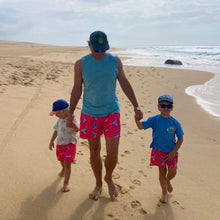 Load image into Gallery viewer, A person walks on a sandy beach with two children, all wearing Breazies' matching Kids Swim Shorts - Lilac Breasted Roller in Coral. The adult partners theirs with a blue tank top under a partly cloudy sky as waves gently meet the shore.