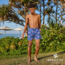 Load image into Gallery viewer, A man stands barefoot on grass by the ocean, wearing Breazies Adult Swim Shorts - Skip Jacks in navy with red patterns. Sun umbrellas and lush greenery surround him under a bright sun. "BREAZIES" is written in the corner.