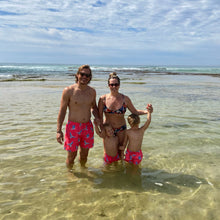 Load image into Gallery viewer, A family stands in clear ocean water on a sunny day. The man and boy sport luxury boys swimwear, wearing Breazies' "Lilac Breasted Roller" coral shorts with matching patterns. The woman complements them with a similar bikini as the child playfully clings to her under the partly cloudy sky.