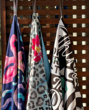 Load image into Gallery viewer, Three colorful microfiber towels hang on wooden hooks: the Beach Towel - Flamingo Queen from Bobums with pink flamingos on a quick-drying blue background, a floral towel with leopard spots, and another featuring abstract black and white graphics with turquoise accents.