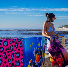 Load image into Gallery viewer, A woman in a bikini top and printed skirt stands by seaside railing on a sunny day, wearing sunglasses as she looks over her shoulder at the calm sea. Nearby, vibrant Bobums Beach Towels in Rainbow Hues Sunset hang, their quick-drying fabric catching rays under the sunny sky.