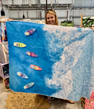 Load image into Gallery viewer, A person holds up a Bobums Beach Towel - Boat View, showcasing its recycled quick-dry material. The towel features vibrant blue ocean waves with an aerial view of boats and white foam below, set against a backdrop of charming shop decorations.