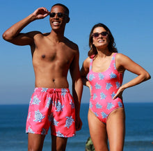 Load image into Gallery viewer, A man and woman smile on a sunny beach, both wearing Breazies' Adult Swim Shorts - Coral Turtles, featuring vibrant blue turtle prints. His quick-drying shorts and her stylish one-piece glisten against the ocean backdrop as they enjoy their sunlit day.