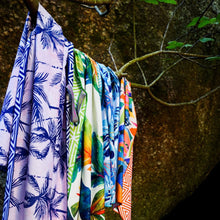 Load image into Gallery viewer, The Bobums Quick Dry Beach Towel - Pink & Black Palms, showcasing tropical prints with palm trees and abstract patterns in vibrant blue, green, orange, and pink hues, hangs drying against a rock backdrop.