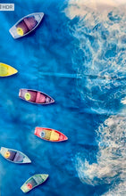 Load image into Gallery viewer, Bobums' Beach Towel - Boat View features an aerial scene of colorful boats on a vivid blue ocean, resembling a painter's palette. The boats have sails crafted from recycled materials in red, yellow, and blue hues with white waves crashing on the right.