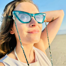 Load image into Gallery viewer, A woman with long hair wears blue cat-eye sunglasses featuring the Sichri Eyewear strap - Livia Beige. She gazes upward, smiling slightly under a clear blue sky while standing outdoors, possibly on a beach with sand visible in the background.