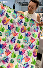 Load image into Gallery viewer, Someone holds a vibrant "Beach Towel - Patterned Pines" by Bobums, showcasing bright greens, pinks, blues, and purples with pineapple patterns. The setting looks like an indoor store or studio.