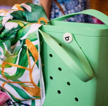 Load image into Gallery viewer, A light green tote bag made from recycled plastics with holes and a "b" button sits next to the Bobums Beach Towel - Green Leaf Jungle. Someone in blue patterned clothing is partly visible in the background.
