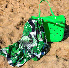 Load image into Gallery viewer, The "Beach Day Ready" tote by Thandana & Bobums, in bright green with circular cutouts and sturdy handles, rests on the sand. Its water-resistant design is perfect for seaside adventures, paired with a quick-dry towel featuring a black, white, and green leaf pattern.