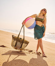Load image into Gallery viewer, On a sunny beach, a woman with sunglasses carries a Thandana Mesh Beach Bag and a large beach ball. She wears a green bikini top and a teal sarong while the waves gently caress the sandy shore in the background.