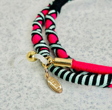 Load image into Gallery viewer, A close-up of Sichri's Rhapsody Hot Pink eyewear strap shows its colorful handcrafted braid with pink, black, and white strands. It features a small gold clasp and charm with engraved letters, resting on a textured light-colored surface.