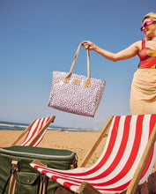 Load image into Gallery viewer, A woman in sunglasses stands on a sandy beach, holding the Thandana Large Beach Bag Laminated Fabric - Seeds White. Two red-and-white striped beach chairs and a green suitcase are in front of her under a clear blue sky.