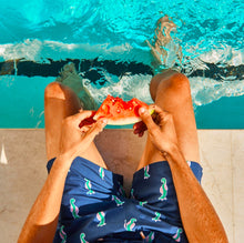Load image into Gallery viewer, Wearing Breazies Adult Swim Shorts in navy sea horse print, a person sits poolside with a watermelon slice. Sunlight creates bright reflections on the water, enhancing the summery and relaxed vibe.