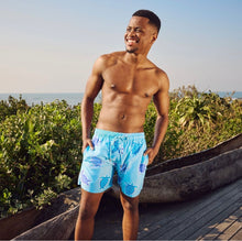 Load image into Gallery viewer, A smiling man stands shirtless on a boardwalk by the ocean, wearing Breazies Adult Swim Shorts - Turtles in Baby Blue. Greenery and waves appear in the background beneath a clear sky.