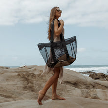 Load image into Gallery viewer, A woman in a beige cover-up and sunglasses walks barefoot on rocky ocean terrain, carrying her Thandana Large Black Mesh Beach Bag. The water-resistant bag protects her belongings as clouds drift in the sky and waves crash nearby.