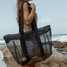 Load image into Gallery viewer, On a rocky beach, someone holds a Large Black Mesh Beach Bag by Thandana, with a leopard print waterproof bag inside. Made from coated mesh to withstand the elements, the bag stands out against the stunning ocean and sky backdrop.