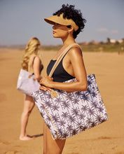 Load image into Gallery viewer, Two women stand on a sandy beach. The woman in front carries the Thandana Large Beach Bag Laminated Fabric - Palms Neutral, while the other in a light-colored swimsuit has a similar waterproof Thandana beach bag in the background.