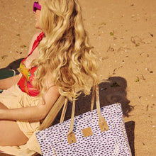 Load image into Gallery viewer, A woman with long, wavy blonde hair relaxes on a beach chair in a red and yellow swimsuit top, cream skirt, and purple sunglasses. Next to her is the Thandana Large Beach Bag Laminated Fabric - Seeds White on the sand.