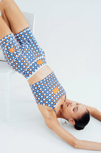 Load image into Gallery viewer, A woman lies on her back on a white chair, legs up and head down, wearing Koko Active's Shorty Shorts - The Court with an athletic top in a blue, orange, and white geometric pattern. The plain white background enhances the vibrant design.