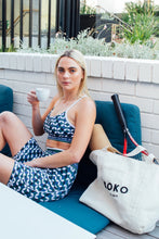 Load image into Gallery viewer, A person with long blonde hair sits on a blue cushioned bench, holding a coffee cup. They're in Koko Active's Retro Grade Shorty Shorts with UV50 protection and have a white tote exposing a tennis racket handle next to them against a white brick wall backdrop.