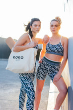 Load image into Gallery viewer, Two women pose confidently against a wall outdoors, wearing matching Shorty Shorts - The Retro Grade by Koko Active, with a leafy-pattern design. One holds a yoga mat and tote bag featuring "Koko," as they stand in bright natural sunlight.