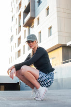 Load image into Gallery viewer, A person crouches outdoors wearing a gray cap, Koko Active's black long-sleeve shirt with UV50 protection, Shorty Shorts in The Retro Grade print, and white high-top sneakers. A modern building with large windows looms in the background.