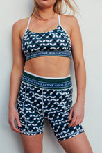 Load image into Gallery viewer, A person sports the "Shorty Shorts - The Retro Grade" by Koko Active, featuring a geometric black, white, and blue pattern with matching sports bra. Ethically designed with UV50 protection, this outfit pops against a plain white background.