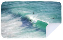 Load image into Gallery viewer, Aerial view of three surfers in the ocean, one riding a large wave while the other two float on their surfboards nearby. The water is a vibrant blue-green with waves creating white foamy trails. In the bottom-right corner, a flipped-up edge reveals a 100% recycled Bobums Extra Large Towel - JBay draped along the shore.