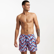 Load image into Gallery viewer, A smiling, shirtless man with short dark hair models Breazies Matching Retro Swimwear trunks in blue, pink, and red wavy stripes made from recycled polyester. He has a left forearm tattoo and stands against a plain background looking left.