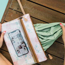 Load image into Gallery viewer, A person with red nail polish holds a green cloth next to a Thandana Sticky Fingers Clear Pouch Laminated Fabric - Palms Natural on a wooden surface, securely containing a smartphone showing images on a social media page.