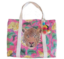 Load image into Gallery viewer, The Beach Bag - Boho Taj by Natural Selection features a vibrant leopard face on poly canvas, with a pink background, green leaves, and yellow flowers. Perfect for beach outings, it includes beige handles, a multicolored pom-pom accessory, and has a convenient waterproof lining.