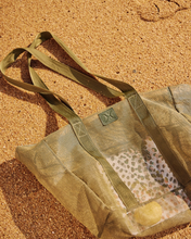Load image into Gallery viewer, A Large Olive Mesh Beach Bag from Thandana lies on golden sand. The water-resistant fabric is partially transparent, revealing a waterproof pocket inside.