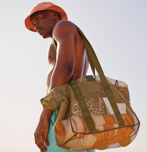 Load image into Gallery viewer, A person stands against a clear sky wearing a coral bucket hat and teal shorts, carrying the Thandana Large Olive Mesh Beach Bag  filled with an orange and white striped towel. The person gazes thoughtfully into the distance.