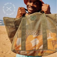 Load image into Gallery viewer, A person on a sandy beach smiles and holds up the Thandana Large Olive Mesh Beach Bag filled with towels. The bag, made from water-resistant fabric, features wide green handles and a logo. The person is wearing a pink hat, and text on the left reads "NEW IN" in a circular pattern.