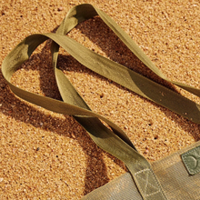Load image into Gallery viewer, A Large Olive Mesh Beach Bag from Thandana rests on a sandy surface, partially buried in the golden grains of sand.