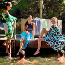 Load image into Gallery viewer, Three girls wrapped in Bobums Extra Large Double Sided Beach Towels - smiling and chatting on a wooden dock by a body of water, while two boys swim nearby. The dock is surrounded by greenery, and sunlight illuminates the scene, creating a relaxed and cheerful atmosphere.