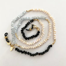 Load image into Gallery viewer, The "Crystal Joy Black" eyewear strap by Sichri, crafted with small, irregular beads in black, gold, white, and pale blue hues, shimmers like crystal on a white surface. Featuring a gold clasp and charm at one end, it resembles an elegant coiled necklace.
