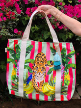 Load image into Gallery viewer, Someone holding the  Lush Leopard Jungle Beach Bag by Natural Selection featuring a leopard surrounded by green banana leaves, bananas and a blue parrot on the left. Pink vertical stripes provide a lively backdrop. It's crafted with durable poly canvas and comfortable cotton straps.