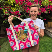 Load image into Gallery viewer, Marzelle is holding the new Amy Taj Beach Bag by Natural Selection. This bag features a leopard's head on a vibrant pink backdrop with teal and pink floral patterns. The beige handles and blue tassels give it a fun yet beachy look. Perfect for a day at the beach.