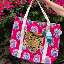 Load image into Gallery viewer, Lady holding the Beach Bag - Amy Taj from Natural Selection by its beige handles. It features a leopard's head on a vibrant pink backdrop with teal and pink floral patterns.