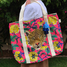 Load image into Gallery viewer, Lady holding a Natural Selection Beach Bag over her shoulder. The Boho Taj print showcases a vibrant pink background with yellow flowers and green leaves, all featuring an intricately detailed leopard face. This bold, colourful design would make a lovely gift.