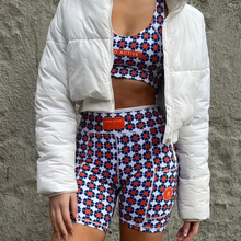 Load image into Gallery viewer, A person wears Koko Active's chic white puffer jacket over a matching sports bra and The Court shorty shorts. The ensemble, adorned with orange and blue flowers on a crisp white background, showcases UV50 protection fabric for style and functionality.