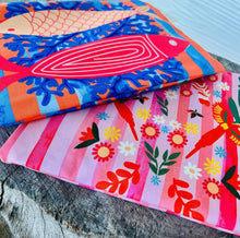 Load image into Gallery viewer, Two wet bags by Natural Selection on a wooden feature. The bags are waterproof with bright and colourful prints and come with a matching beach wrap/sarong.