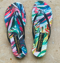 Load image into Gallery viewer, Vibrant flip-flops featuring red, blue, green, and yellow abstract swirls with contrasting black straps. The "Treasure Beach Made in USA" branded soles add comfort while resting on a light brown surface. These stylish sandals are known as Flip Flops - Tropical Black by Treasure Beach.