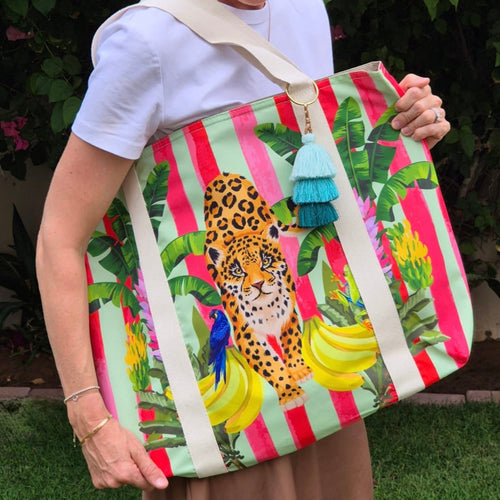 A lady holding the  Lush Leopard Jungle Beach Bag by Natural Selection featuring a leopard surrounded by green banana leaves, bananas and a blue parrot on the left. It's crafted with durable poly canvas and comfortable cotton straps plus a key chain with tassels.