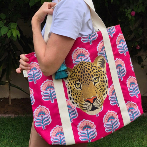 Lady holding the Amy Taj beach bag over her shoulders using the beige cotton straps. This bag is by Natural Selection featuring a leopard's head on a vibrant pink backdrop with teal and pink floral patterns. Made from durable material with waterproof lining and a cute blue tassel added for decoration.